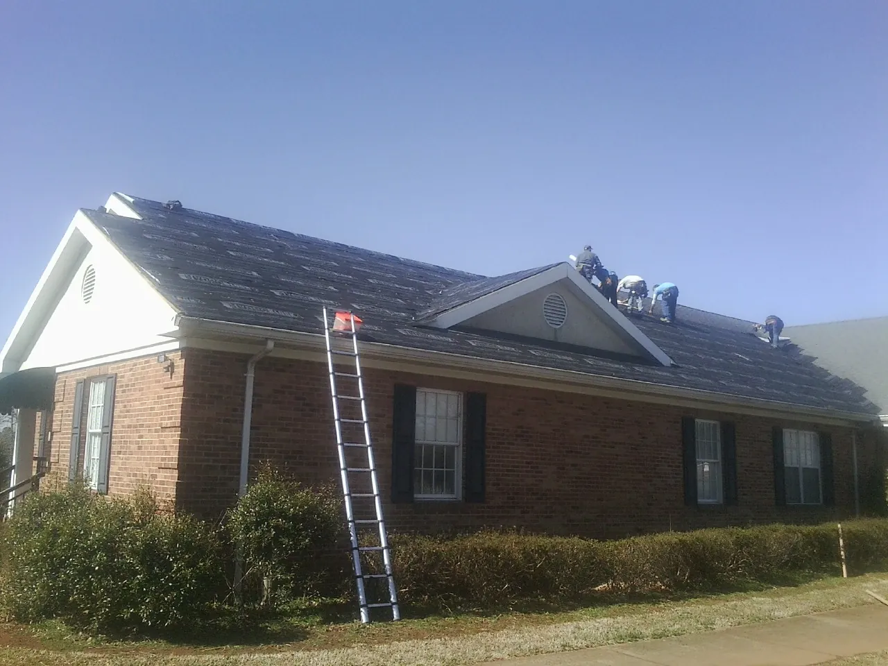 Crew tearing shingles off a brick ranch roof with a ladder set — Emergency Roof Repair work by our crew in Pipestone