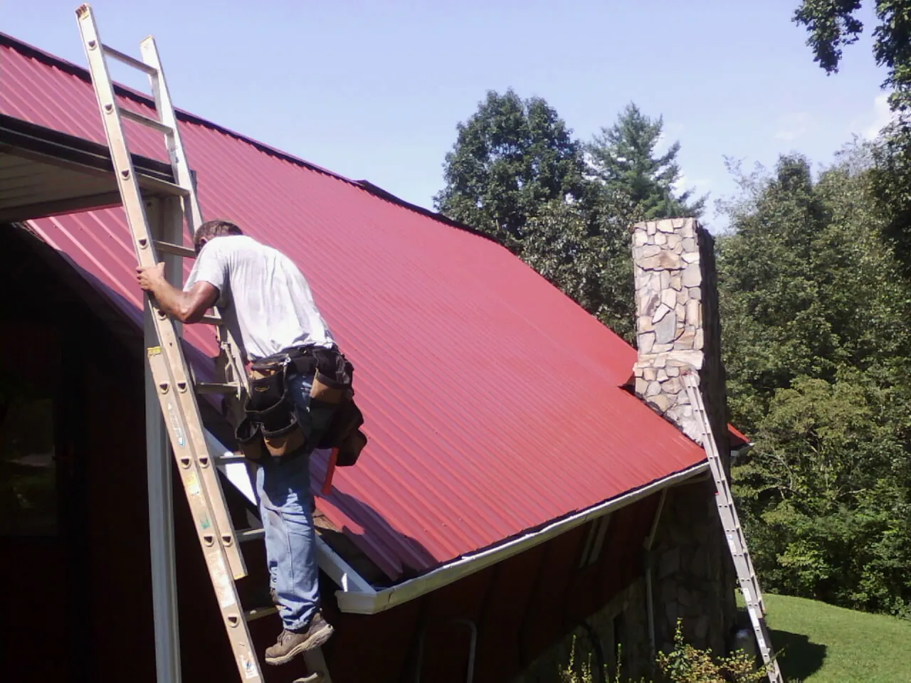 Roofer on a ladder beside a red metal roof and stone chimney — Soffit & Fascia Repair by Roofing Brothers in Pipestone
